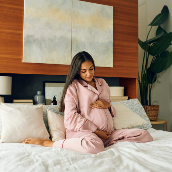 Trudnica u ružičastom ogrtaču, sjedi na krevetu i miluje svoj trbuh.