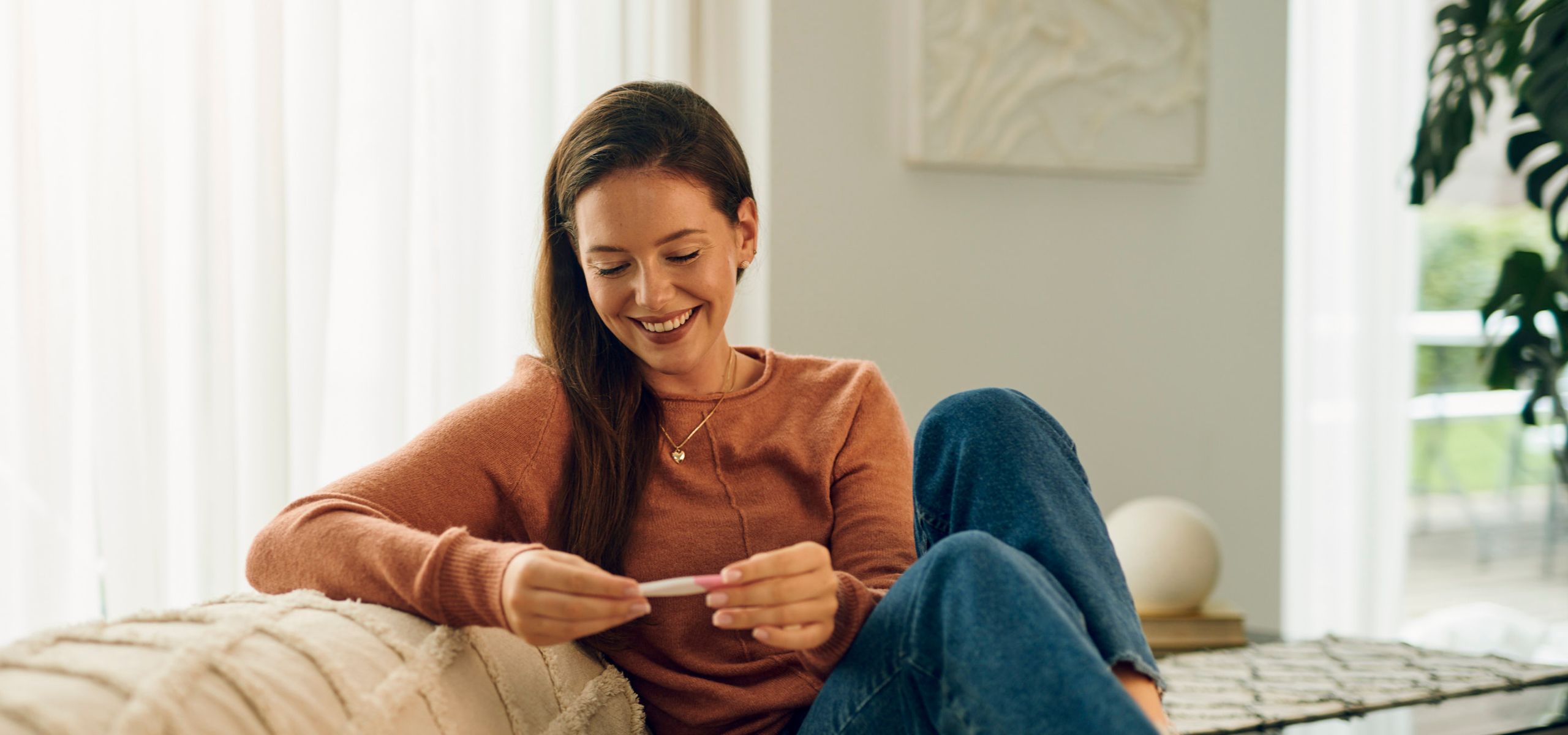 Trudnica sjedi na kauču, gledajući pozitivan test na trudnoću, planira budućnost.