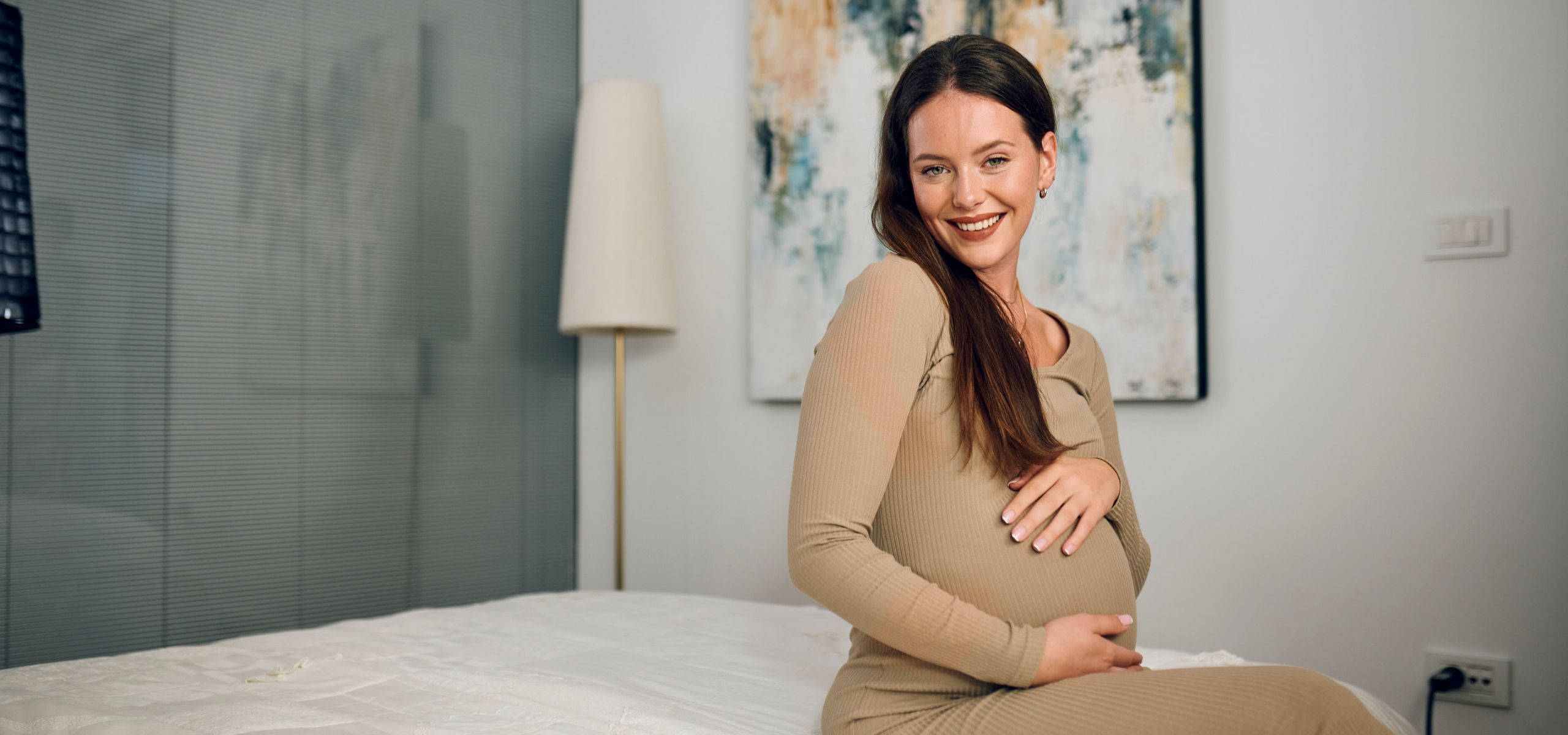 Trudnica sjedi na krevetu, miluje svoj trbuh – trenutak povezivanja s bebom.