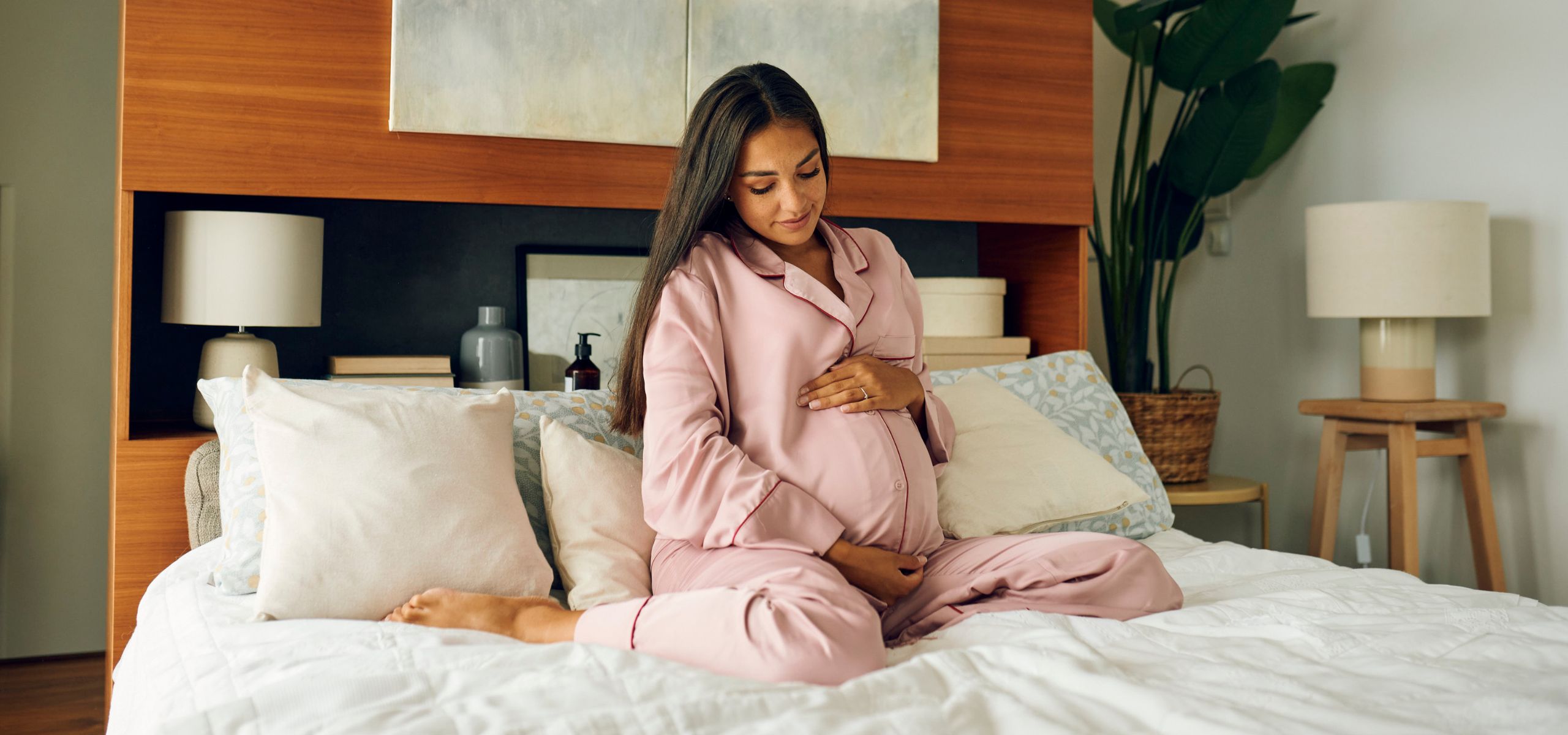 Trudnica u ružičastom ogrtaču, sjedi na krevetu i miluje svoj trbuh.