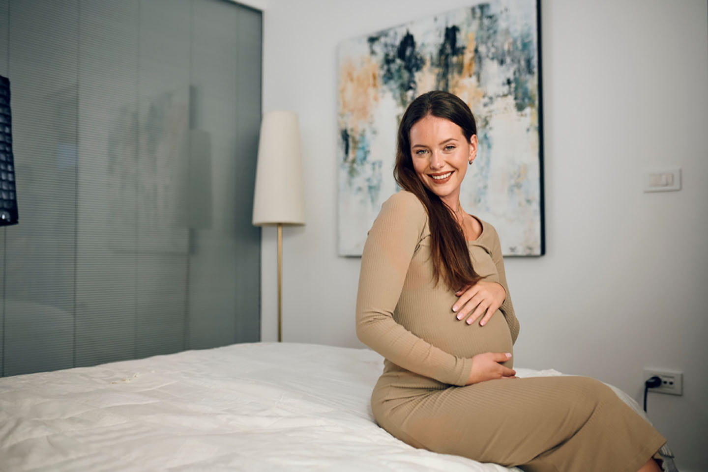 Trudna žena smiješi se dok sjedi na krevetu – sretan trenutak tijekom trudnoće.