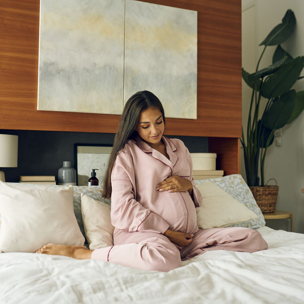 Trudnica u ružičastom ogrtaču sjedi na krevetu i drži se za trbuh.